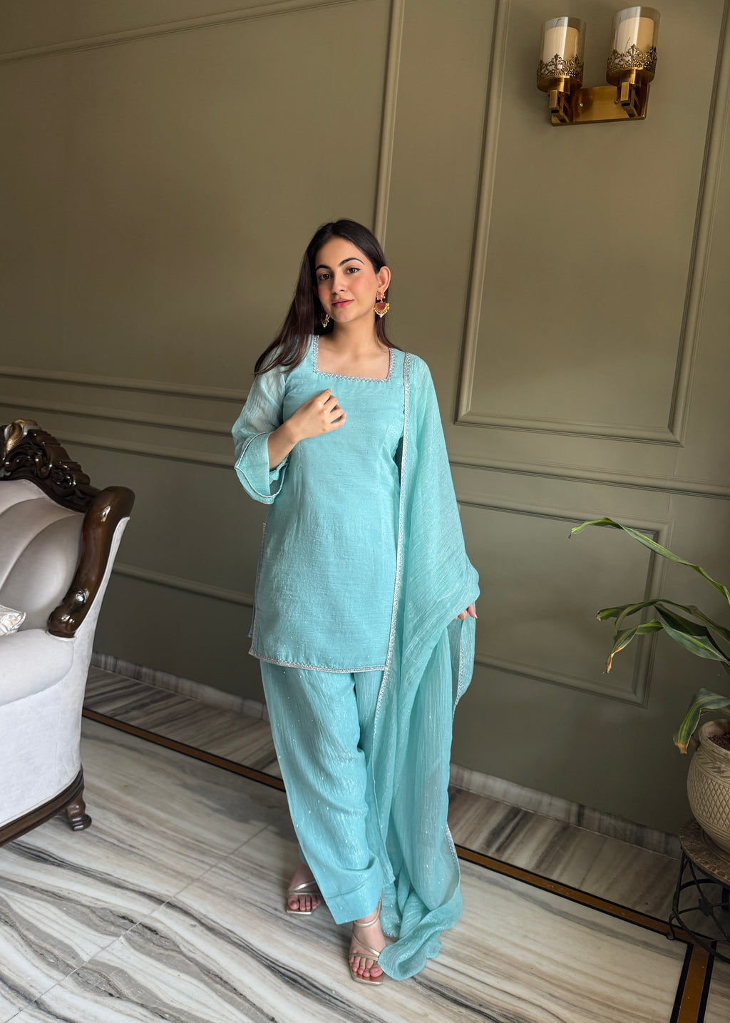 Woman in a light blue traditional outfit standing in a room with a neutral wall and white chair.